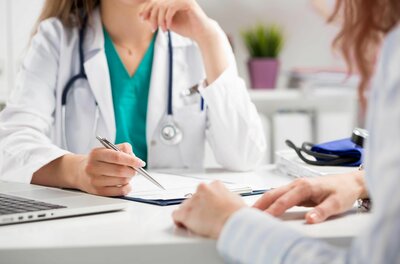 Ärztin im weißen Kittel mit Stethoskop führt ein Beratungsgespräch mit einer Patientin an einem Schreibtisch.