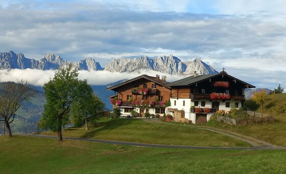 Zwei traditionelle Holzhäuser auf einer Wiese vor einem schroffen Gebirgsmassiv mit Wolkenband.