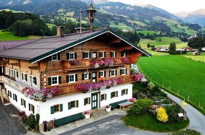 Traditionelles Alpenhaus aus Holz und Stein mit Blumenschmuck an den Balkonen vor grünen Wiesen und Bergen.