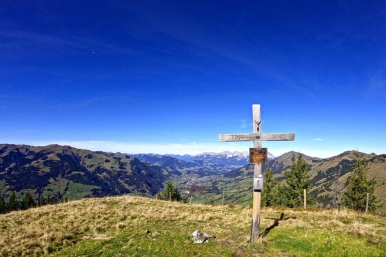 Holzernes Gipfelkreuz auf einer grasbewachsenen Bergkuppe vor weitem Talpanorama und blauem Himmel.