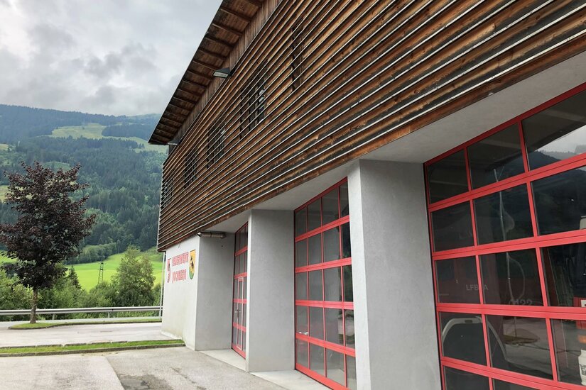 <p>Das Feuerwehrgebäude von Jochberg mit Holzverkleidung und großen Fenstern. Im Hintergrund sind grüne Wiesen und Berge zu sehen.</p>
