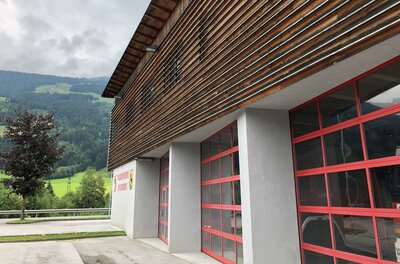 <p>Das Feuerwehrgebäude von Jochberg mit Holzverkleidung und großen Fenstern. Im Hintergrund sind grüne Wiesen und Berge zu sehen.</p>