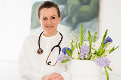 Dr. Laura Gelny Eine freundliche Ärztin mit einem Stethoskop um den Hals lächelt in die Kamera. Der Hintergrund ist hell und modern gestaltet.