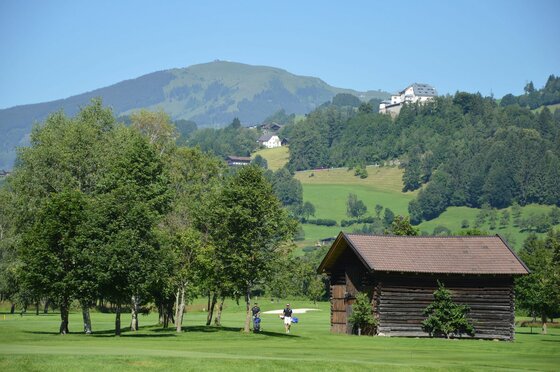 Golfplatz mit Holzstadel, Bäumen und zwei Personen vor bewaldeten Hügeln und Bergen.