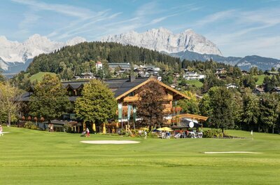 Ein Holzgebäude am Rand eines Golfplatzes vor bewaldeten Hügeln und schroffen Berggipfeln.