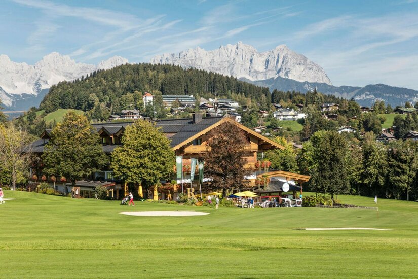 Ein Holzgebäude am Rand eines Golfplatzes vor bewaldeten Hügeln und schroffen Berggipfeln.