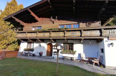 Ansicht Front App. Barbara Zweistöckiges alpines Wohnhaus mit weißer Fassade im Erdgeschoss und dunklem Holzbalkon im Obergeschoss.
