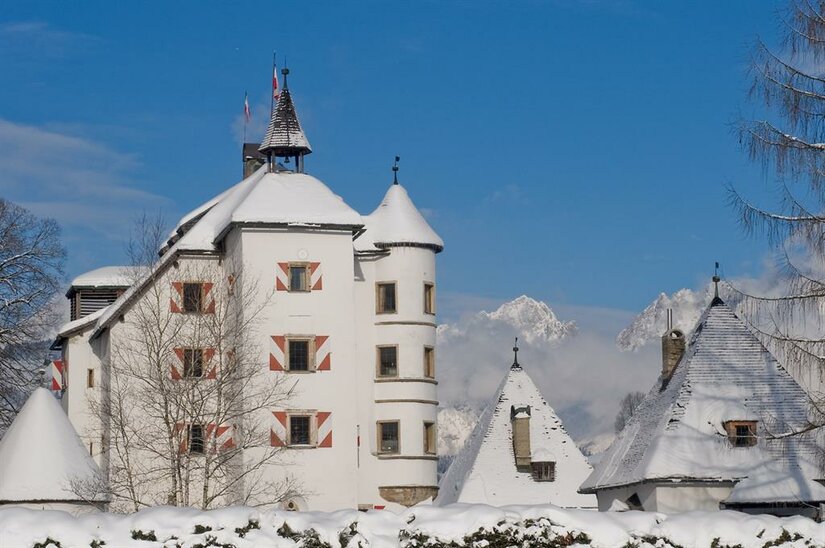 Winter am Schloss Münichau Weißes Schloss mit schneebedeckten Dächern und rot-weiß gestreiften Fensterläden vor Gebirgskulisse.