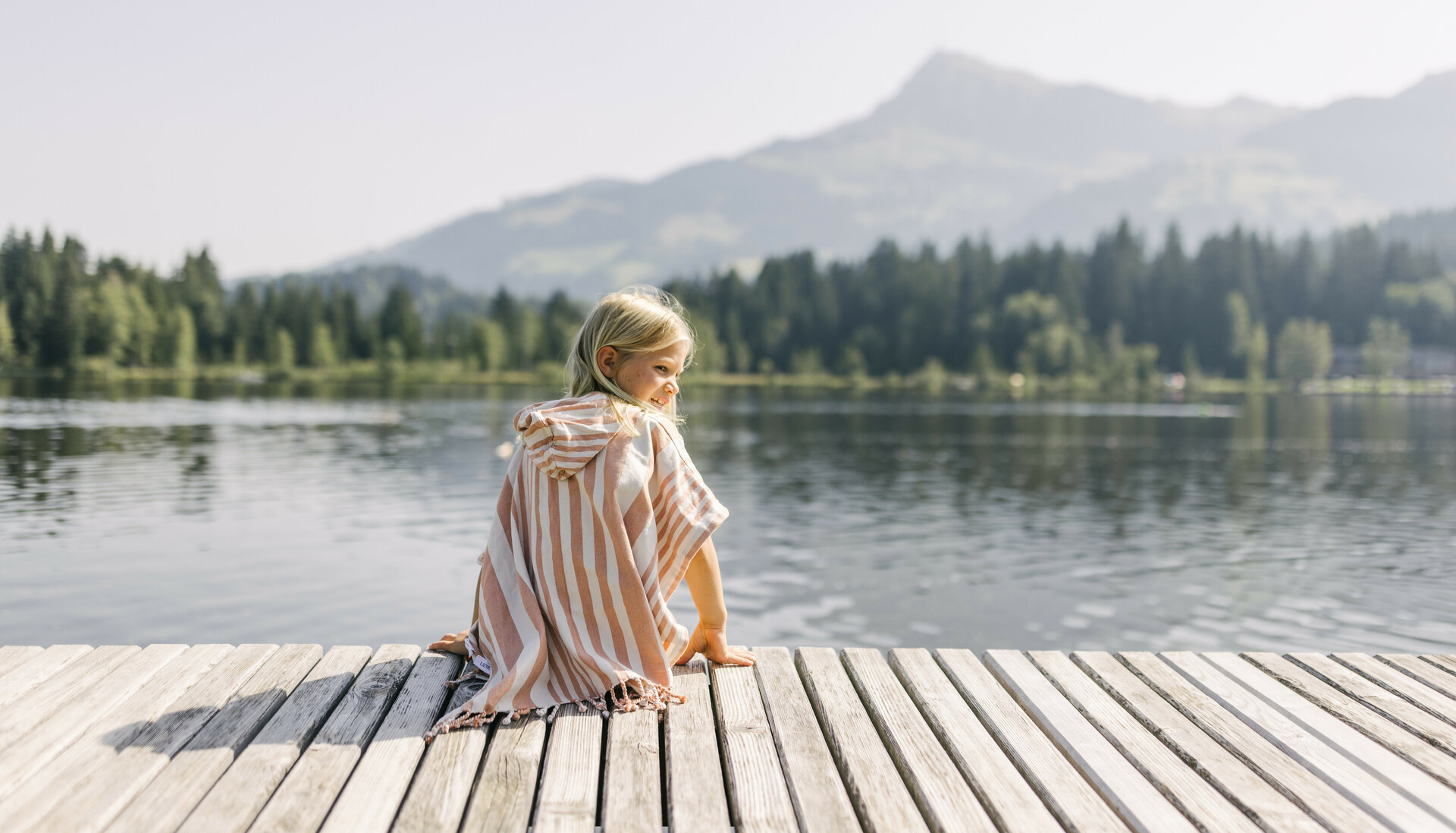 Ein kleines Mädchen sitzt am Ufer des Schwarzsees in Kitzbühel, umgeben von Bergen und Bäumen, und schaut lächelnd über das Wasser.