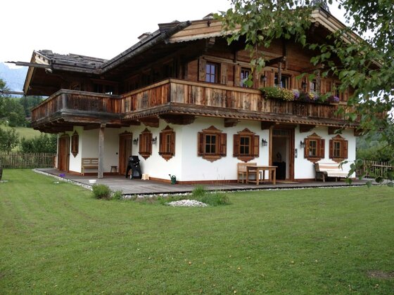 Zweistöckiges Holzchalet mit weißem Erdgeschoss, Holzbalkon und grüner Rasenfläche.