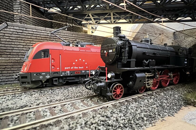 Modelleisenbahn Kitzbüheler Alpen Modelle einer roten Elektrolokomotive und einer schwarzen Dampflokomotive auf Schienen unter einer Brücke.