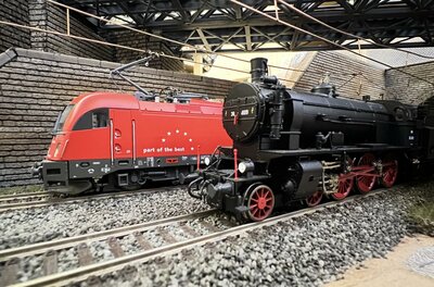 Modelleisenbahn Kitzbüheler Alpen Modelle einer roten Elektrolokomotive und einer schwarzen Dampflokomotive auf Schienen unter einer Brücke.