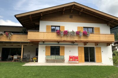 Zweistöckiges alpines Wohnhaus mit Holzfassade, Balkonblumen und einer Terrasse mit Sitzgelegenheiten.