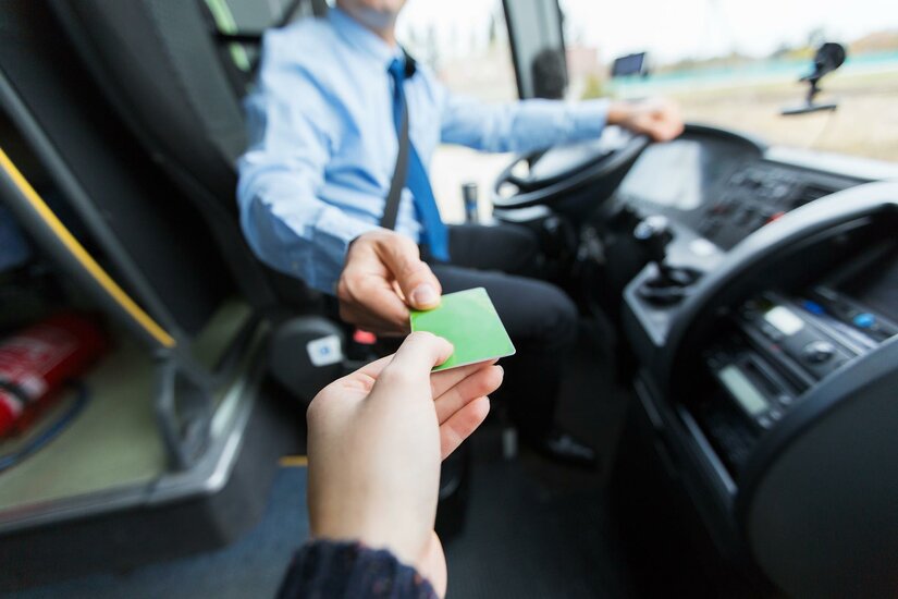Ein Fahrgast gibt dem Busfahrer eine grüne Fahrkarte. Der Fahrer sitzt am Steuer des Busses.