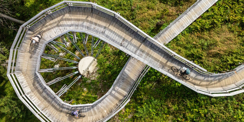 In Kitzbühel fahren zwei Radfahrer über eine geschwungene Holzbrücke in grüner, bewaldeter Umgebung.