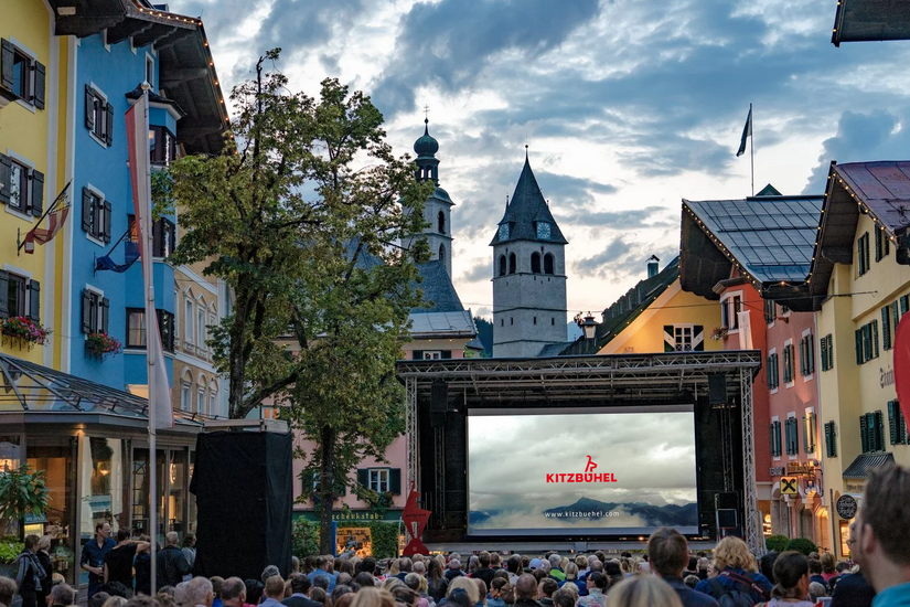 Eine große Menschenmenge versammelt sich auf einem Platz in Kitzbühel, um einen Film auf einer Leinwand zu sehen.