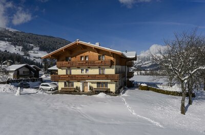 Dreistöckiges alpines Gästehaus mit Holzbalkonen in einer verschneiten Winterlandschaft unter blauem Himmel.