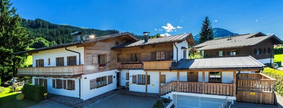 Zweistöckiges Landhaus mit Holzfassade, Balkonen und Satteldach vor bewaldeten Bergen.