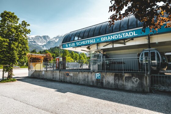 Talstation Bergbahn Scheffau Eine Seilbahnstation in der Bergregion mit Blick auf die verschneiten Berge. Die Umgebung ist grün und einladend, ideal für Ausflüge in der Natur.