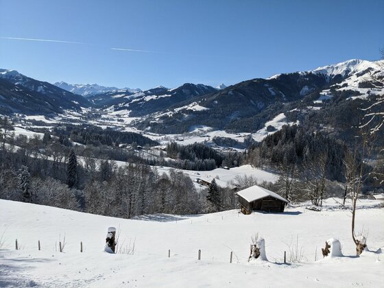 Winterlandschaft bei Oberaigen Verschneite Berglandschaft mit einer Holzhütte im Vordergrund und einem weiten Tal unter blauem Himmel.