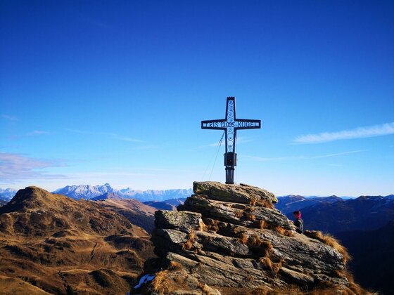 Gipfelkreuz der 5-Gipfel-Tour Ein metallisches Gipfelkreuz auf einem felsigen Berggipfel vor blauem Himmel.