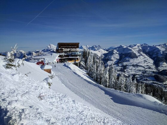 Berghütte an einer Skipiste in verschneiter Gebirgslandschaft unter blauem Himmel.