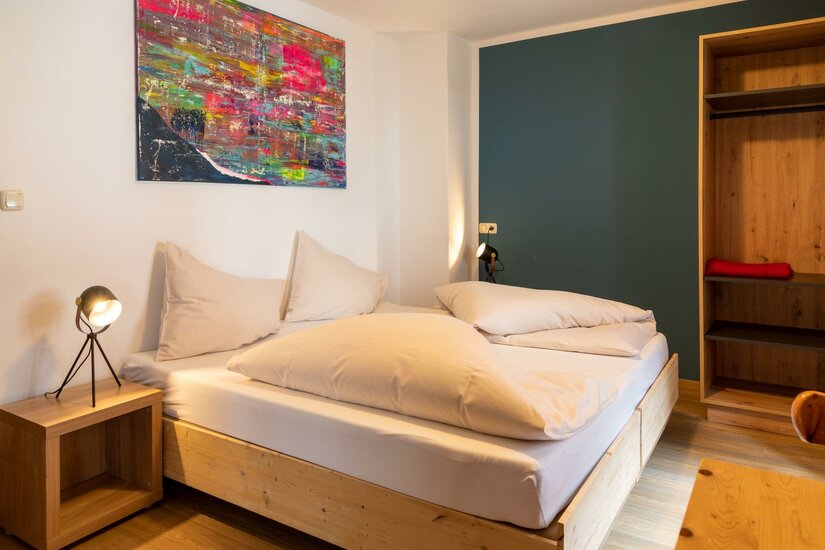 Schlafzimmer im Safestay Kitzbühel Modernes Doppelbett aus Holz mit weißer Bettwäsche in einem hellen Zimmer mit buntem Wandbild.