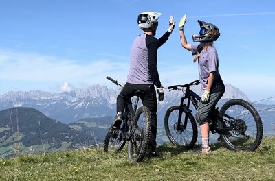 Zwei Mountainbiker geben sich auf einem Berggipfel vor einem Alpenpanorama ein High-Five.