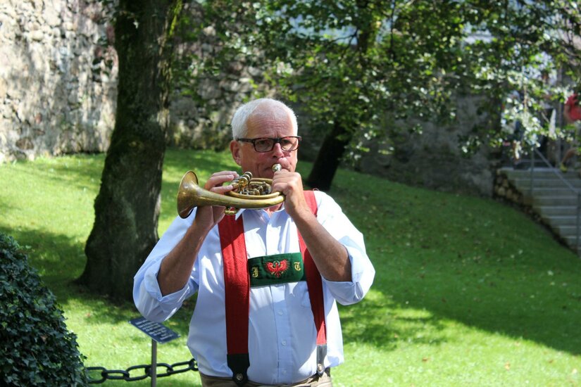 Ein älterer Mann spielt ein Instrument in einem grünen Park. Er trägt eine traditionelle Tracht mit Hosenträgern.