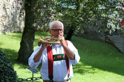 Ein älterer Mann spielt ein Instrument in einem grünen Park. Er trägt eine traditionelle Tracht mit Hosenträgern.