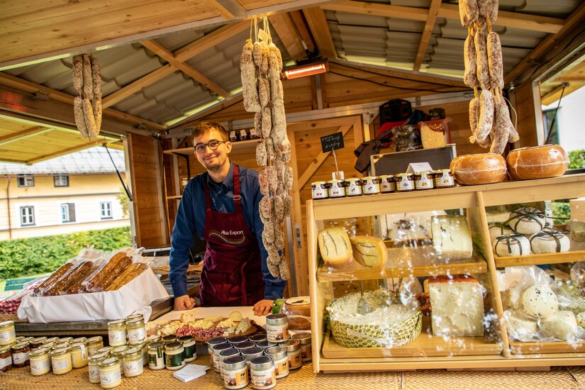 Ein Verkäufer in einer Holzhütte präsentiert Käse, Salami und Gläser mit regionalen Spezialitäten.
