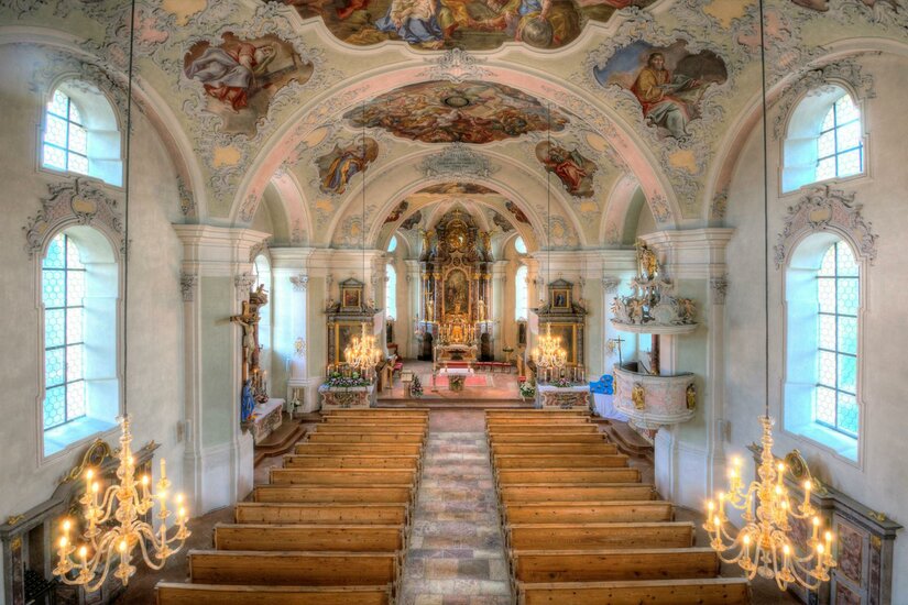 Pfarrkirche "Zum Heiligen Wolfgang" Eine wunderschöne Kircheninneneinrichtung mit gewölbter Decke und kunstvollen Fresken. Das Interieur ist hell erleuchtet und zeigt Holzbänke sowie goldene Kronleuchter.