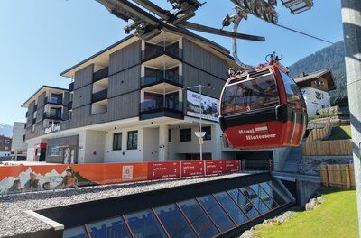 Eine moderne Gondelbahn transportiert Passagiere zu einem Gebirgshotel. Im Hintergrund sind die Berge und der blaue Himmel zu sehen.