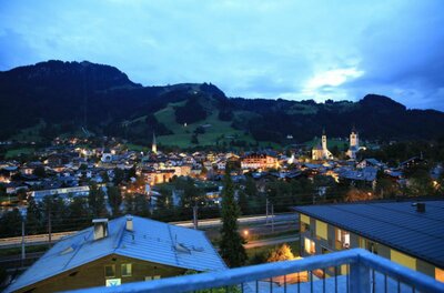Blick von einer Terrasse auf die beleuchtete Stadt Kitzbühel bei Dämmerung vor bewaldeten Bergen.