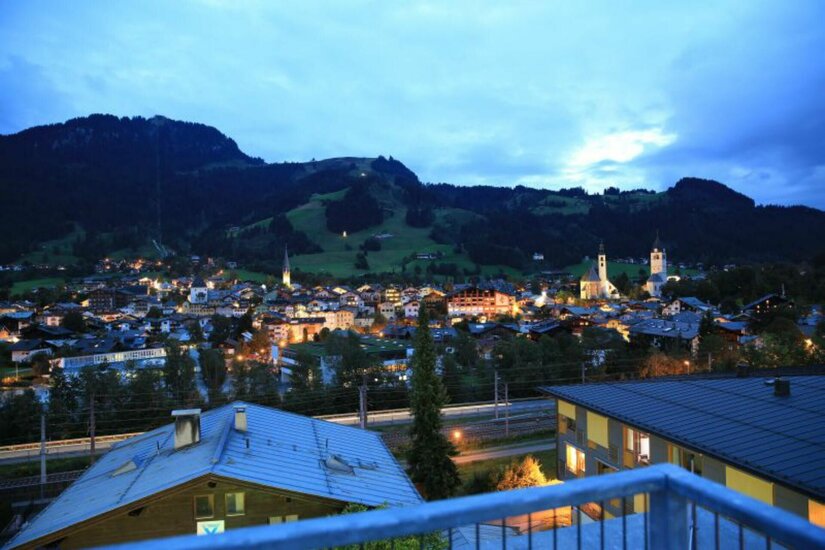 Blick von einer Terrasse auf die beleuchtete Stadt Kitzbühel bei Dämmerung vor bewaldeten Bergen.
