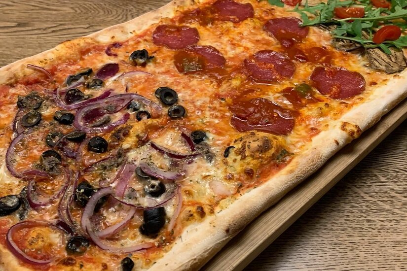 Längliche Pizza mit verschiedenen Belägen wie Salami, Oliven, roten Zwiebeln und Rucola auf einem Holzbrett.