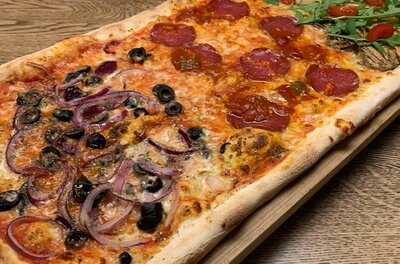 Längliche Pizza mit verschiedenen Belägen wie Salami, Oliven, roten Zwiebeln und Rucola auf einem Holzbrett.