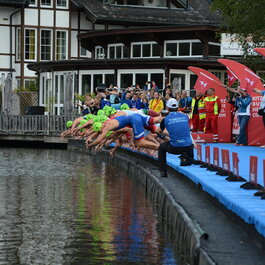 Athleten in grünen Schwimmkappen springen gleichzeitig ins Wasser, während Zuschauer am Ufer jubeln und filmen.