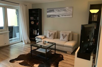 Wohnzimmer mit hellem Sofa, Glastisch, Kuhfellteppich und einem Bergbild an der Wand.