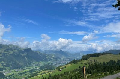 Aussicht von der Branderalm Blick von einem Schotterweg auf ein grünes Tal mit bewaldeten Hängen und Bergen unter blauem Wolkenhimmel.