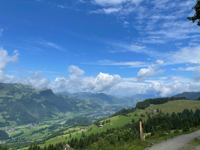 Blick von einem Schotterweg auf ein grünes Tal mit bewaldeten Hängen und Bergen unter blauem Wolkenhimmel.
