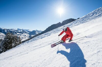 Skifahrer in roter Kleidung fährt eine präparierte Piste bei Sonnenschein hinunter.