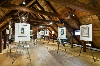 Ein Ausstellungsraum mit mehreren Kunstwerken auf Ständern. Die Holzdecke und der Boden verleihen dem Raum eine gemütliche Atmosphäre.