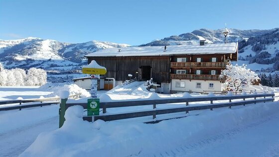 Ansicht Winter Angererhof Verschneiter Bauernhof mit Holzfassade und gelbem Wegweiser vor schneebedeckten Bergen.