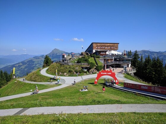 Berghaus an der Panoramarunde Berghaus auf einem Gipfel mit befestigten Wanderwegen und einem roten Torbogen unter blauem Himmel.