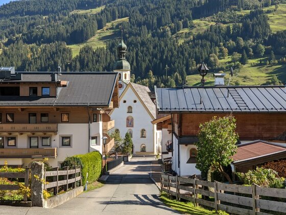 Jochberger Dorfansicht mit Kirche Blick durch eine Dorfstraße auf eine weiße Kirche mit Zwiebelturm vor bewaldetem Berghang.