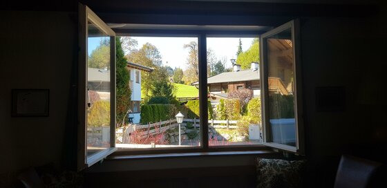 Blick durch ein geöffnetes Fenster auf benachbarte Häuser, Gärten und eine grüne Wiese unter blauem Himmel.
