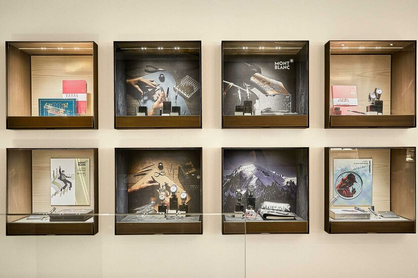 Montblanc Boutique Vitrinen Acht quadratische Glasvitrinen an einer hellen Wand mit Schreibgeräten, Uhren und Markenmotiven von Montblanc.