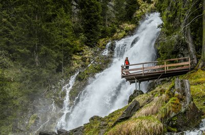 Ein beeindruckender Wasserfall umgeben von grünen Bäumen. Eine Person steht auf einer Holzplattform und genießt die Aussicht.