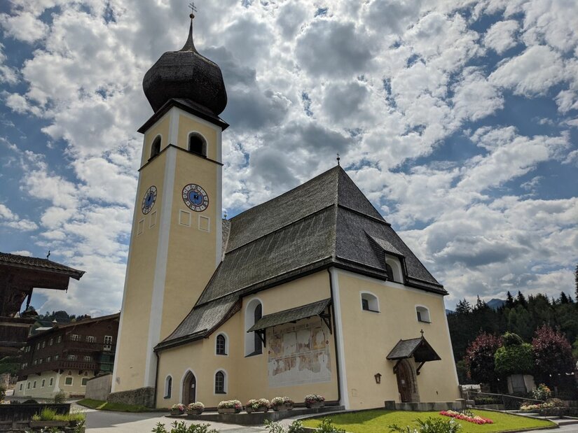 Pfarrkirche in Aurach mit markantem Zwiebelturm und Schindeldach vor bewölktem Himmel.